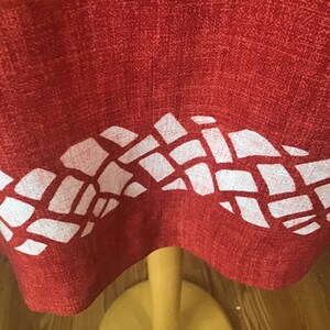 May include: Close-up of a red fabric with a white geometric pattern along the bottom edge. The fabric appears to be a tablecloth or similar textile, with a textured surface. The pattern is made up of irregular white shapes.