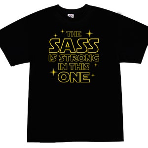 May include: Black t-shirt with a yellow graphic that says "The Sass Is Strong In This One" with stars around the text.