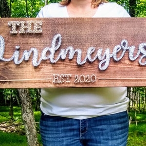Può includere: Un cartello di legno con il testo "The Lindmeyers Est 2020" creato con l'arte della corda. Il cartello è tenuto da una persona davanti a uno sfondo verde.