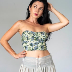 May include: A woman wearing a strapless floral print top with a light gray skirt. The top has a sweetheart neckline and a fitted bodice. The skirt is gathered at the waist and has a ruffled hem.