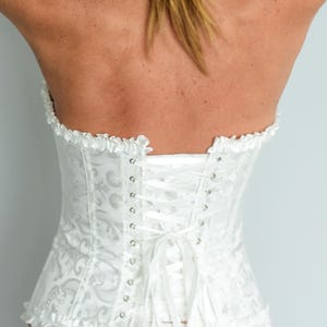 May include: A white corset with a floral pattern and a lace-up back. The corset has a ruffled trim along the top and bottom edges. The back is laced with white satin ribbon.