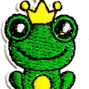 May include: A green frog wearing a yellow crown. The frog has big eyes and a smiling mouth. The frog is sitting on its bottom with its legs tucked in.