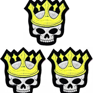 Könnte beinhalten: Drei gestickte Totenkopf-Patches in Schwarzweiß mit gelben Kronen. Die Totenköpfe haben schwarze Augen und ein zahniges Grinsen.