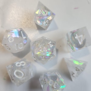 Puede incluir: Un conjunto de siete dados transparentes e iridiscentes con numeración blanca. Los dados tienen varias formas, incluyendo un d4, d6, d8, d10, d12 y d20. Los dados tienen un efecto brillante de arcoíris y están dispersos sobre una superficie blanca.