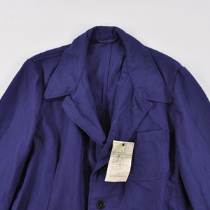 Op de afbeelding: Een donkerblauw werkjasje met knoopsluiting en een enkele zak op de linkerborst. Het jasje heeft een label aan de zak.