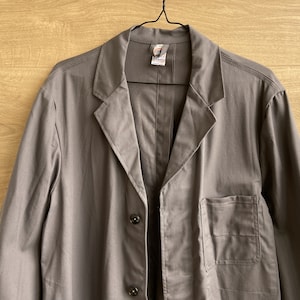 May include: A gray work coat with a notched lapel, three buttons, and a chest pocket. The coat is hanging on a black hanger against a light brown wooden background. The coat has a tag on the inside collar.