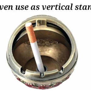 May include: A round, antique-style ashtray in bronze tones, with a cigarette inserted. The ashtray has a decorative floral pattern around the base and the text "Even use as vertical stand" in black.