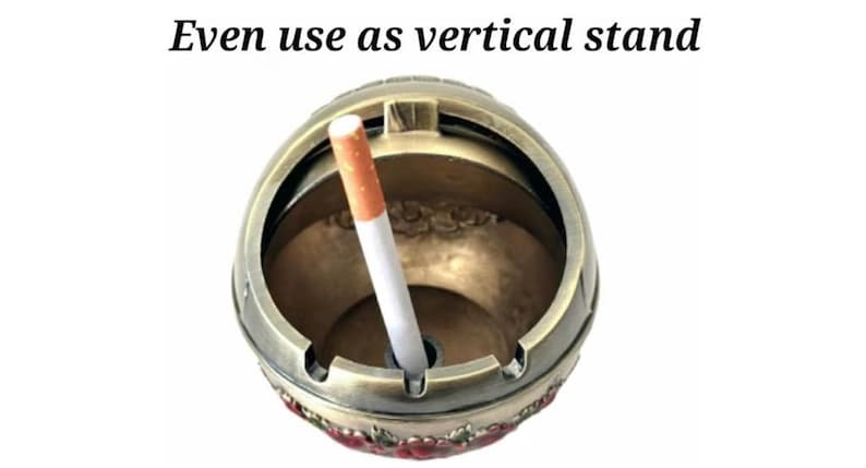 May include: A round, antique-style ashtray in bronze tones, with a cigarette inserted. The ashtray has a decorative floral pattern around the base and the text "Even use as vertical stand" in black.