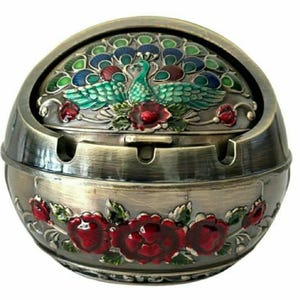 Puede incluir: Un cenicero decorativo de metal con tapa abisagrada. La tapa presenta un diseño de pavo real con detalles en azul, verde y rojo. La base está adornada con detalles de rosas rojas. El cenicero tiene un acabado bronce.