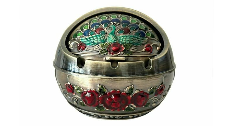 May include: A decorative metal ashtray with a hinged lid. The lid features a peacock design with blue, green, and red accents. The base is adorned with red rose details. The ashtray has a bronze finish.