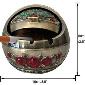 May include: An antique-style ashtray, 3.5 inches tall and 3.9 inches wide, with a hinged lid and a cigarette. The ashtray is bronze-colored with red rose detailing and green accents. The ashtray is round and has a decorative floral design.