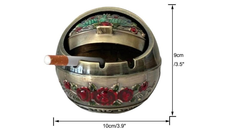 May include: An antique-style ashtray, 3.5 inches tall and 3.9 inches wide, with a hinged lid and a cigarette. The ashtray is bronze-colored with red rose detailing and green accents. The ashtray is round and has a decorative floral design.