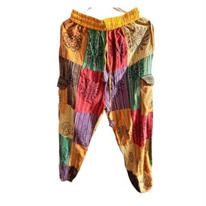 May include: Loose-fitting pants featuring a patchwork design with alternating rectangular panels in yellow, red, green, and purple. The fabric displays black line-art illustrations of suns, flowers, and mushrooms. The pants have an elastic waistband.