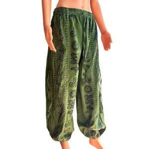 May include: Olive green loose-fitting trousers featuring a black printed pattern of suns, spirals, and floral motifs. The pants have an elasticized waistband with a drawstring tie and gathered cuffs at the ankles, creating a relaxed silhouette.