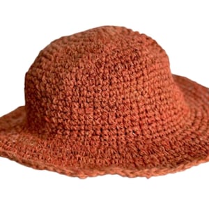 May include: A woven sun hat in a warm orange-brown hue. The hat features a rounded crown and a wide, slightly curved brim. The texture of the hat appears to be made of a natural fiber, possibly raffia or straw, with a visible woven pattern.
