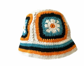 Puede incluir: Un sombrero de cubo blanco de ganchillo con rayas naranjas, negras y azules y un diseño floral.