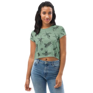 Könnte beinhalten: Hellgrünes Crop-Top mit einem sich wiederholenden Muster aus schwarzen Insektenillustrationen. Das Oberteil hat kurze Ärmel und einen runden Ausschnitt. Das Model trägt eine blaue Jeans.