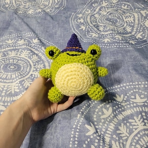 May include: A green crocheted frog wearing a purple witch hat. The frog has a yellow belly and black eyes.