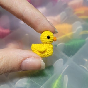 May include: A small yellow crocheted duck with black eyes and an orange beak. The duck is being held by a hand.