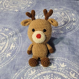 May include: A crocheted reindeer toy with brown fur, a red nose, and brown antlers. The toy is sitting on a blue and white patterned fabric.