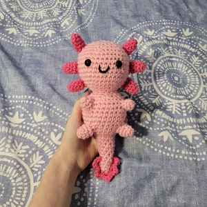 May include: A handmade pink crochet axolotl plushie with dark pink gills and tail. The axolotl has black safety eyes and a smiling mouth. The plushie is approximately 6 inches tall.