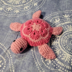 May include: A crocheted pink turtle toy with black eyes. The turtle is lying on a blue and white patterned fabric.