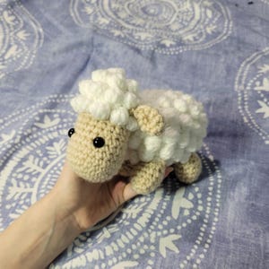 May include: A crocheted white and beige sheep toy with black eyes. The sheep is sitting on a blue and white patterned surface.