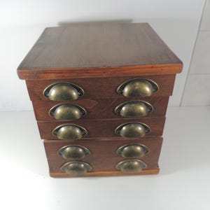 Puede incluir: Un pequeño mueble de cajones de madera con cuatro cajones. El mueble está hecho de madera marrón oscuro y tiene tiradores de color latón. Los cajones están apilados y la parte superior del mueble es plana.