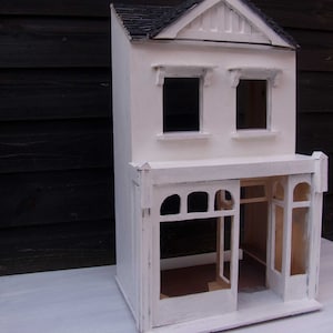 May include: A white doll's house with a shop front, two windows, and a door. The doll's house is made of wood and has a simple design.