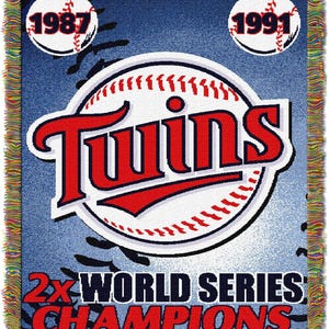 May include: A blue and white woven tapestry with the Minnesota Twins logo and the text "2x World Series Champions". The logo features a baseball with the word "Twins" in red and white letters. The years "1987" and "1991" are displayed in white circles with red outlines.