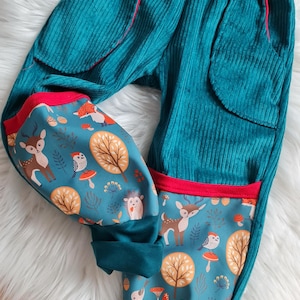 Può includere: Pantaloni in velluto a coste turchese con bordini rossi e una stampa di animali della foresta sulle gambe. La stampa presenta cervi, uccelli, funghi e alberi.