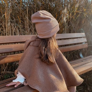 Może przedstawiać: Dziecko w brązowym płaszczu i pasującej czapce, siedzące na drewnianej ławce. Peleryna ma kaptur i opada na ramiona. Czapka jest miękka, dzianinowa. Dziecko ma na sobie jasne legginsy i buty.