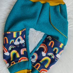 Könnte beinhalten: Türkisfarbene Fleecehose mit senfgelbem Bund und Bündchen. Die Taschen haben einen marineblauen Stoff mit bunten Regenbogen- und Wolkenmustern. Die Hose ist auf Komfort und Stil ausgelegt.