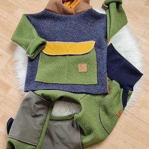 Może przedstawiać: Dwuczęściowy strój dziecięcy w odcieniach niebieskiego, zielonego i brązowego. Bluza z kapturem ma dużą przednią kieszeń i brązowy kaptur z żółtą podszewką. Spodnie są w kolorze oliwkowym z granatowymi mankietami. Małe logo na kieszeni.