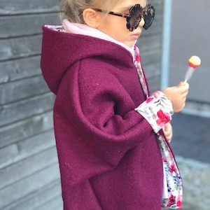 May include: A burgundy hooded cape with a floral print lining. The cape is made of a soft, warm fabric and has a relaxed fit. The child is wearing a pair of brown sunglasses.