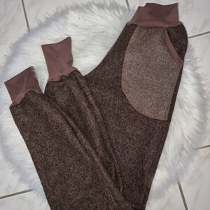 May include: Brown fleece sweatpants with a large patch pocket on the back. The sweatpants have a brown ribbed waistband and cuffs.