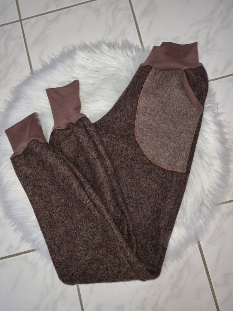 May include: Brown fleece sweatpants with a large patch pocket on the back. The sweatpants have a brown ribbed waistband and cuffs.