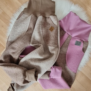 Può includere: Due paia di pantaloni in pile marroni e rosa con una cintura marrone a contrasto. I pantaloni hanno un design a patchwork e una grande tasca sul retro. I pantaloni hanno un'etichetta che dice "Mini Zicke".