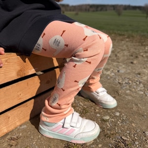 May include: Peach-colored leggings with a white goose pattern and text. The leggings are worn with white sneakers with pink stripes and a black top. The child is sitting on a wooden crate outdoors.