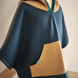 May include: A navy blue hooded poncho with a tan hood and pocket. The hood is trimmed with green fabric. The poncho has a large tan pocket on the front and a small tag on the pocket.