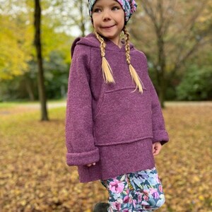 Könnte beinhalten: Ein junges Kind trägt einen lilafarbenen Kapuzenpullover mit einer Mütze mit Blumenmuster. Der Pullover hat eine große Tasche auf der Vorderseite. Das Kind trägt eine Leggings mit Blumenmuster und steht in einem Park mit heruntergefallenen Blättern auf dem Boden.
