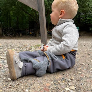 Könnte beinhalten: Ein kleines Kind in grauen, fleecegefütterten Hosen mit gelben Akzenten sitzt auf einem Schotterweg. Das Kind trägt einen grauen Pullover und schaut zur Seite.