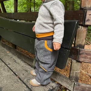 Può includere: Un bambino indossa un maglione grigio e pantaloni grigi e neri con accenti gialli. I pantaloni hanno un pannello di tessuto contrastante grigio e nero sulle gambe.