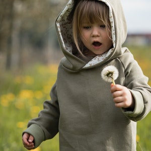 Può includere: Un bambino piccolo che indossa una felpa con cappuccio verde con fodera a stampa floreale. Il bambino tiene un dente di leone in mano e lo guarda.