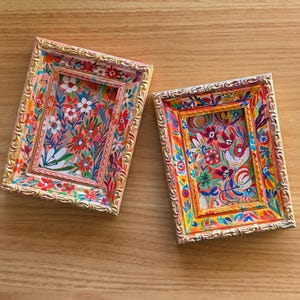 Puede incluir: Dos marcos rectangulares ornamentados con obras de arte florales. Los marcos tienen ribetes dorados y están pintados con colores vibrantes, incluyendo rosa, rojo, naranja y azul. La obra de arte interior presenta un patrón floral colorido.