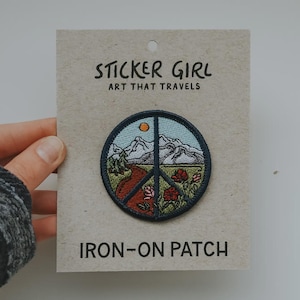 May include: A round iron-on patch with a peace sign design featuring a mountain landscape, a road, and flowers. The patch is on a cardboard backing with the text "STICKER GIRL ART THAT TRAVELS" and "IRON-ON PATCH".