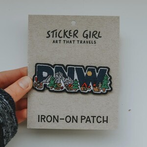 May include: An iron-on patch with the letters "PNW" in dark blue, featuring a mountain range, trees, and a campfire design. The patch is on a cardboard backing with the text "STICKER GIRL ART THAT TRAVELS" and "IRON-ON PATCH".