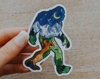 May include: A die-cut sticker in the shape of a Bigfoot silhouette. The interior design features a night sky with a crescent moon, mountains, trees, a river, and a campsite. The sticker is held by a hand.