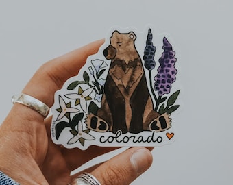 May include: A sticker featuring a watercolor illustration of a brown bear sitting among flowers and purple lupine. The word "colorado" is written in white script below the bear, with a small orange heart. The sticker is held in a hand.