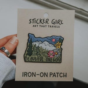 May include: An embroidered iron-on patch on a tan card labeled STICKER GIRL ART THAT TRAVELS. The patch is shaped like Oregon and features a landscape with green pine trees, white mountains, a blue sky, a pink sun, and yellow and pink flowers.
